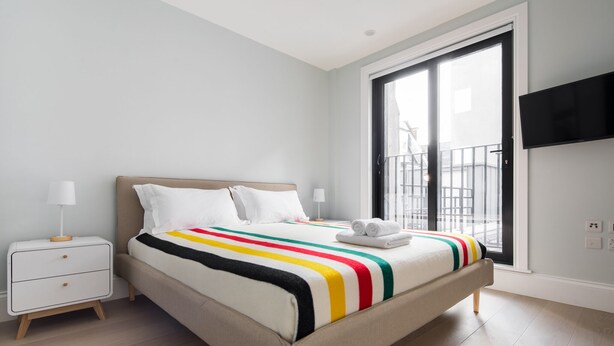 Quarto de hotel moderno com uma cama grande, mesa de cabeceira, televisão na parede e varanda com guarda-corrimão.