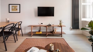 Uma sala de estar moderna com uma mesa de jantar de madeira, cadeiras pretas, uma TV fixada na parede e um radiador.