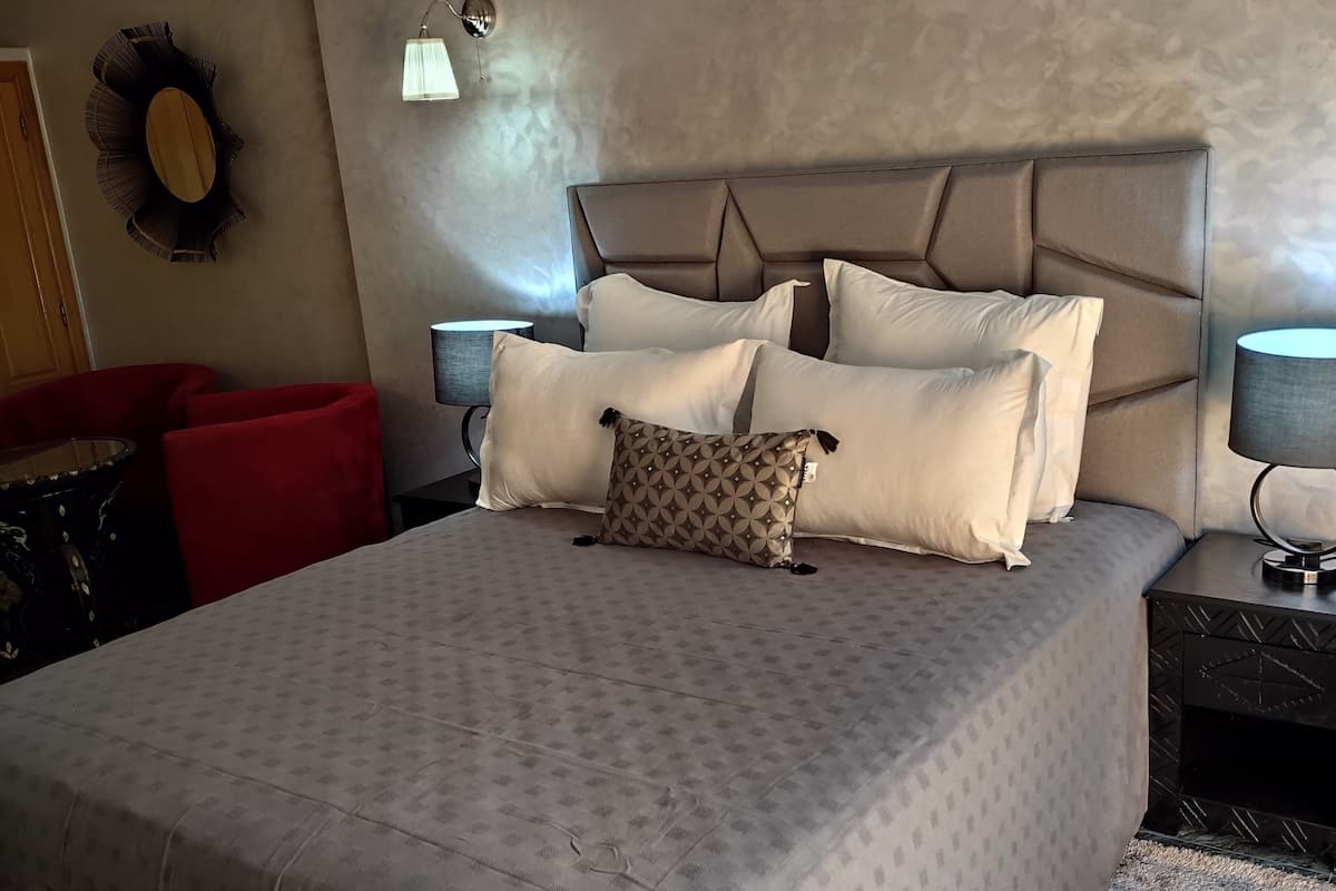 A neatly made bed with a patterned bedspread, a headboard, and multiple pillows. There is a wall-mounted air conditioner above the bed, a small table with a lamp, and a red chair in the corner.