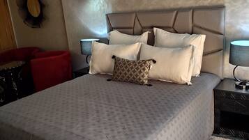 A neatly made bed with a patterned bedspread, a headboard, and multiple pillows. There is a wall-mounted air conditioner above the bed, a small table with a lamp, and a red chair in the corner.