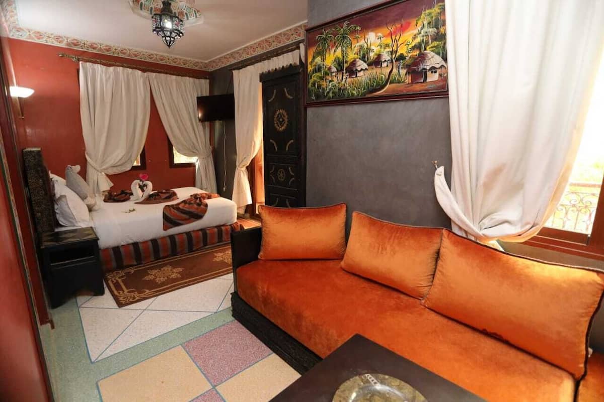 A room with a bed, a sofa, a nightstand, a lamp, and a painting on the wall.
