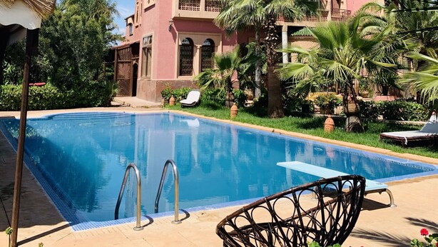 Une piscine entourée de chaises longues, de palmiers et d’un bâtiment rose doté de balcons.