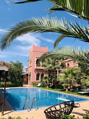 Ein Swimmingpool mit Liegestühlen, umgeben von Palmen und einem rosa Gebäude mit Balkonen.