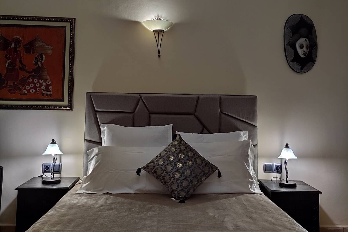 A bedroom with a bed, two bedside tables with lamps, a wall-mounted light fixture, a decorative headboard, and a framed picture on the wall.