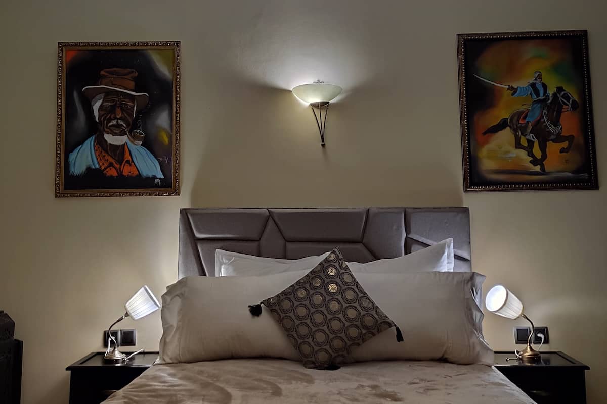 A bedroom with a large bed, two bedside tables, a wall-mounted light, and two framed artworks.