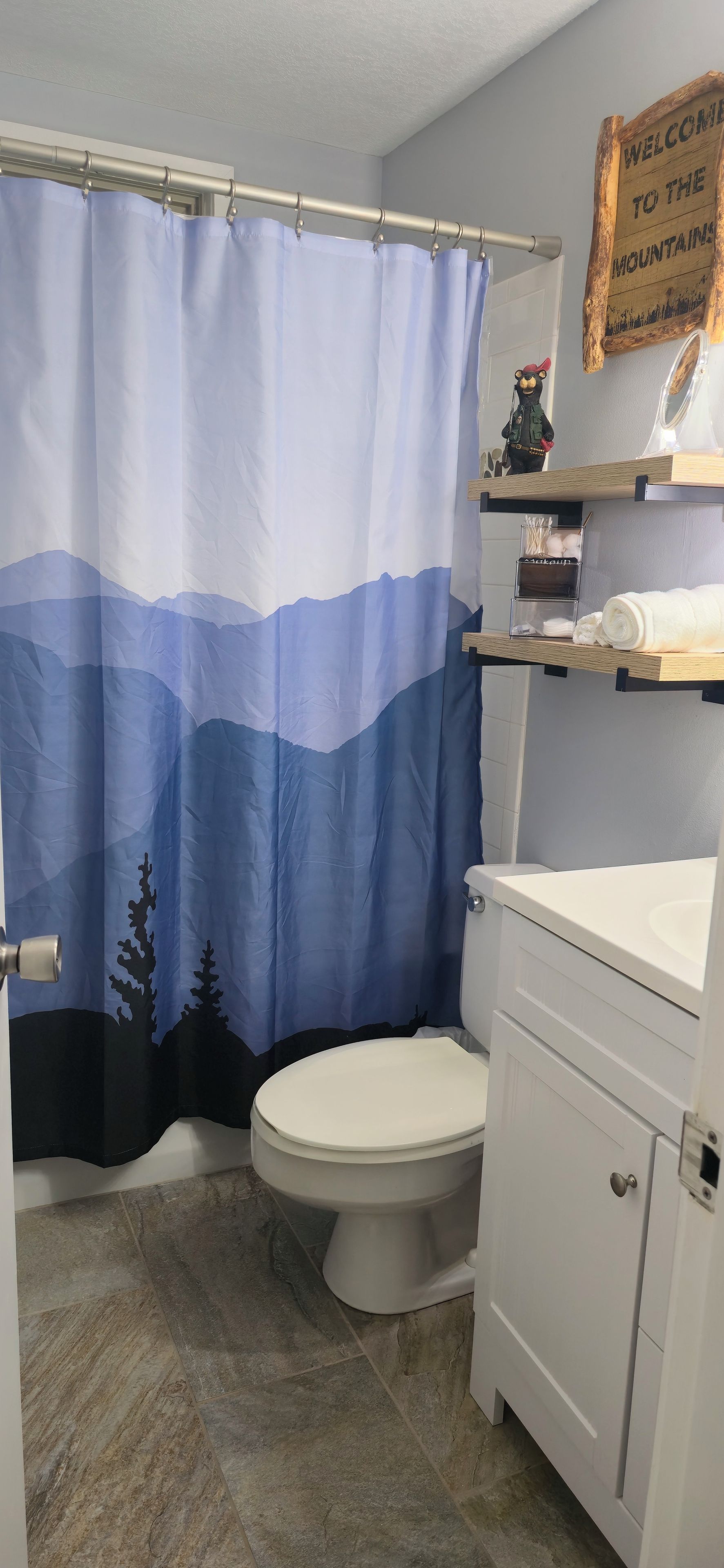 Kylpyhuone, jossa on vuoristomaisemaa kuvaava suihkuverho, wc-istuin ja puinen hylly, jossa on hygieniatuotteita.