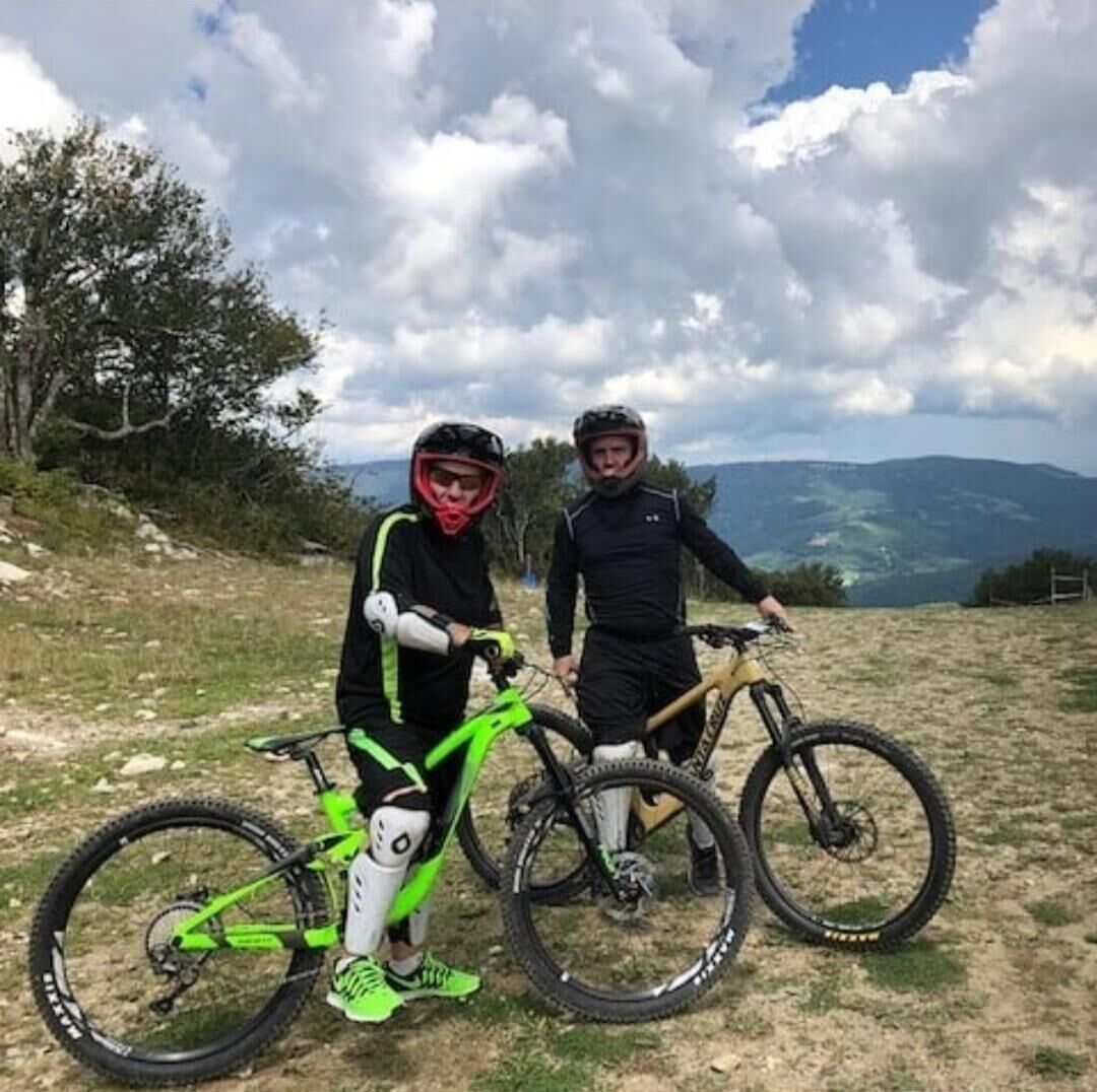 Due persone in piedi su una collina erbosa con biciclette, circondate da alberi e montagne sullo sfondo.