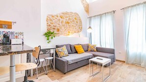 A modern living room with a stone accent wall, a sofa with patterned cushions, a dining area with a coffee machine, and a large window with curtains. - SunCastle Romantic Loft (Alicante/Alacant)
