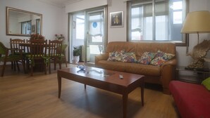 A living room with a brown leather sofa, a wooden coffee table, a dining table with chairs, and a large window with blinds.