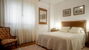 A bedroom with a bed, a chair, a nightstand, a lamp, and a window with curtains.