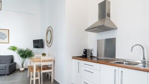 A compact kitchen with white cabinets, a stainless steel sink, and a modern stove. There's a wooden dining table with chairs, a potted plant, and a flat-screen TV mounted on the wall.