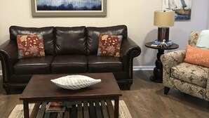 Una sala de estar con un sofá de cuero marrón oscuro, una mesa de centro de madera y un sillón estampado. Hay dos cuadros en la pared y una lámpara sobre una mesita.