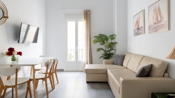A modern living room with a beige sofa, a dining table with chairs, a TV mounted on the wall, and a large window with curtains. - Sanlúcar de Barrameda  6 (Sanlucar de Barrameda)