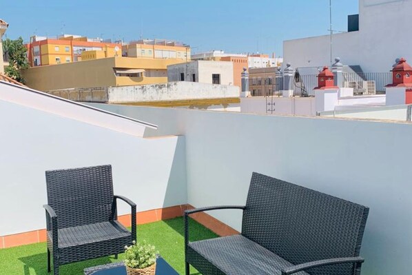 A rooftop terrace with two wicker chairs, a small potted plant, and a view of surrounding buildings.