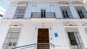 Un edificio blanco con una puerta de madera grande, varias ventanas con barrotes de metal y un balcón con barandilla metálica.