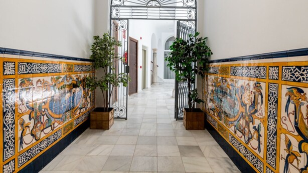 Un pasillo con paredes de azulejos decorativos, suelo de baldosas y una reja metálica que conduce a otra zona.