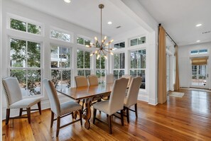 Uma área de jantar com uma mesa de madeira grande e seis cadeiras, janelas amplas com vista para vegetação, e um lustre moderno.