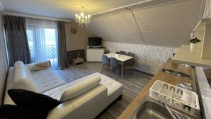 Una habitación de hotel moderna con sofá, mesa de comedor, televisor, pequeña cocina y balcón.