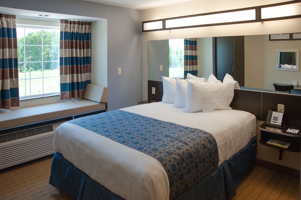 A hotel room with a large bed, a window seat with curtains, a bedside table with a phone, and a mirror.