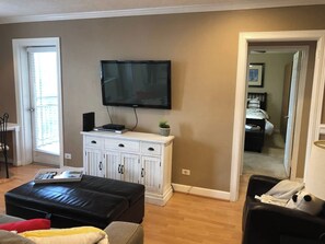 A living room with a flat-screen TV, a white cabinet, a black leather ottoman, and a doorway leading to another room.