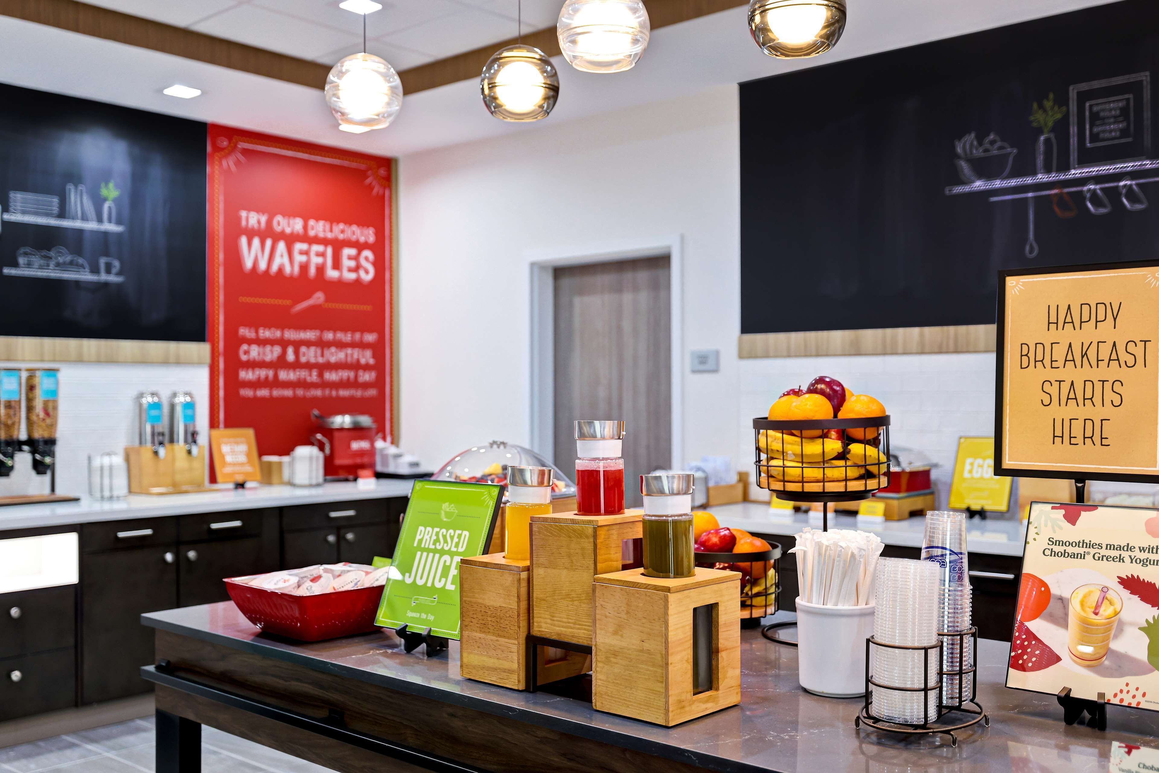 A breakfast buffet area with a red sign advertising waffles, a juice station, and a display of fruits and yogurt.