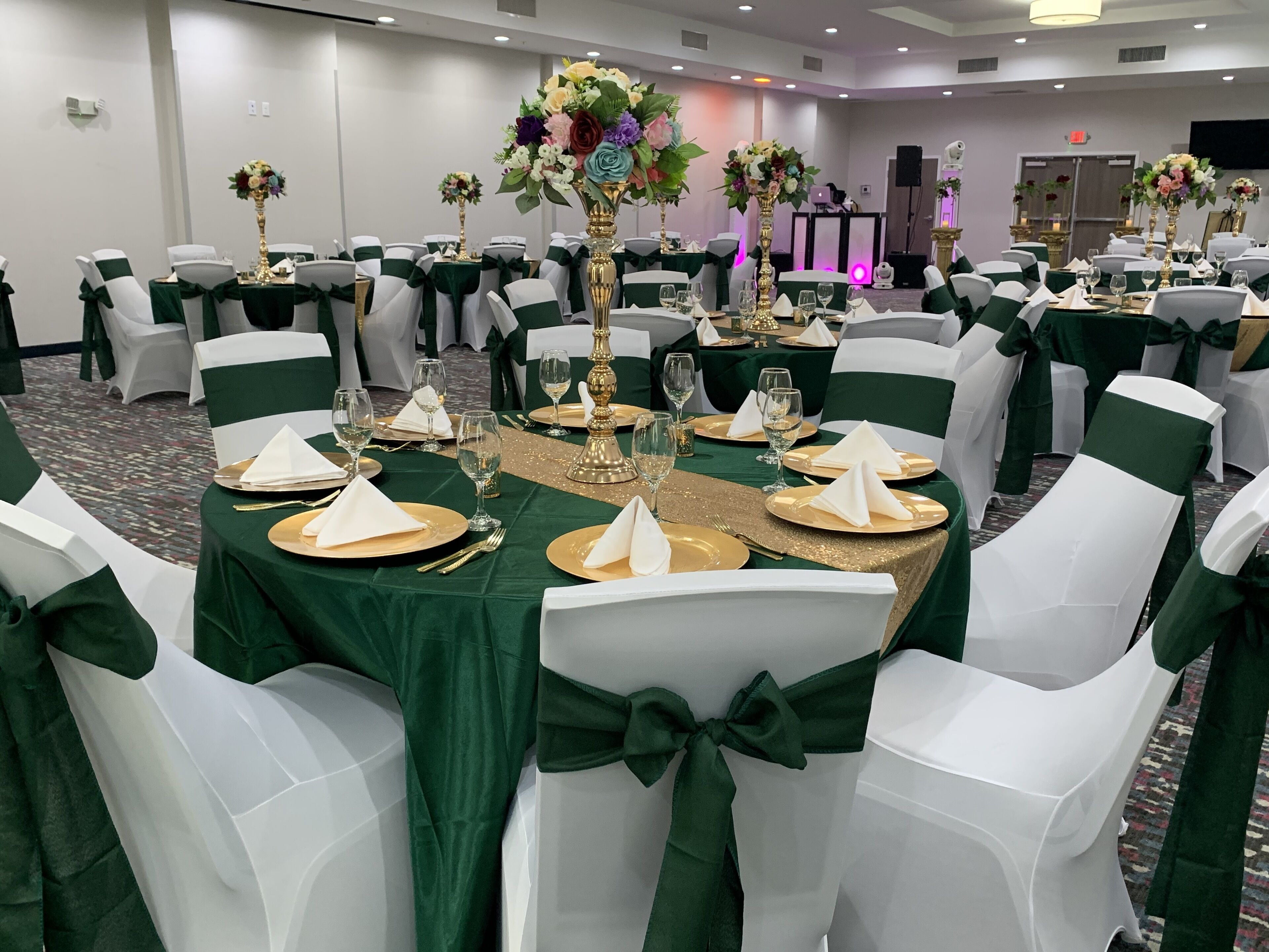 A banquet hall with round tables set for a formal event, featuring green tablecloths, white chairs with green sashes, and gold accents.