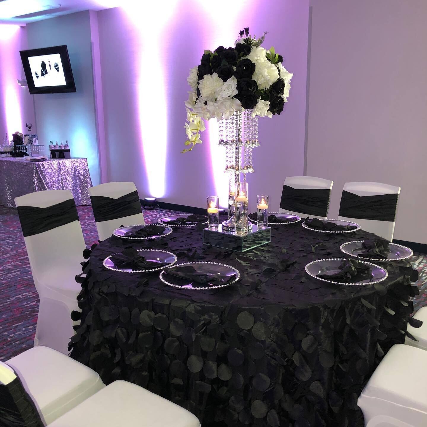 A round table with a black tablecloth, surrounded by white chairs with black sashes. A tall, clear glass centerpiece with flowers is placed in the center.