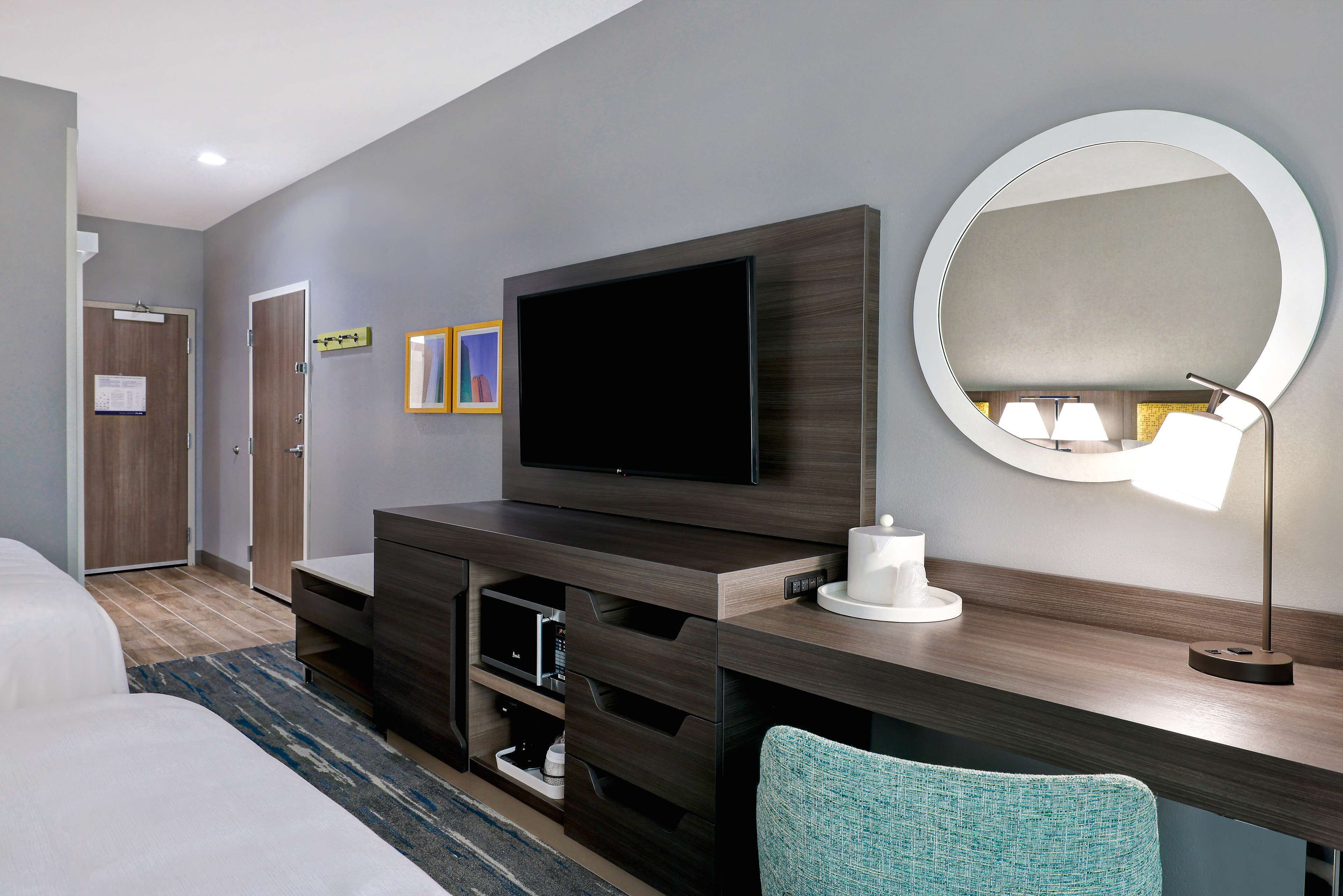 Habitación de hotel moderna con un televisor de pantalla plana grande, un espejo redondo, un escritorio con lámpara y una cama con alfombra azul y gris a rayas.