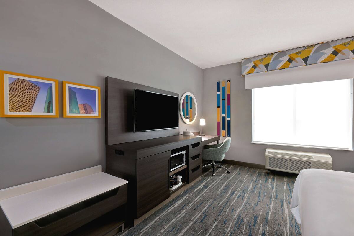 A hotel room with a flat-screen TV, a desk with a chair, a bedside table, a large window with a view, and a wall-mounted mirror.