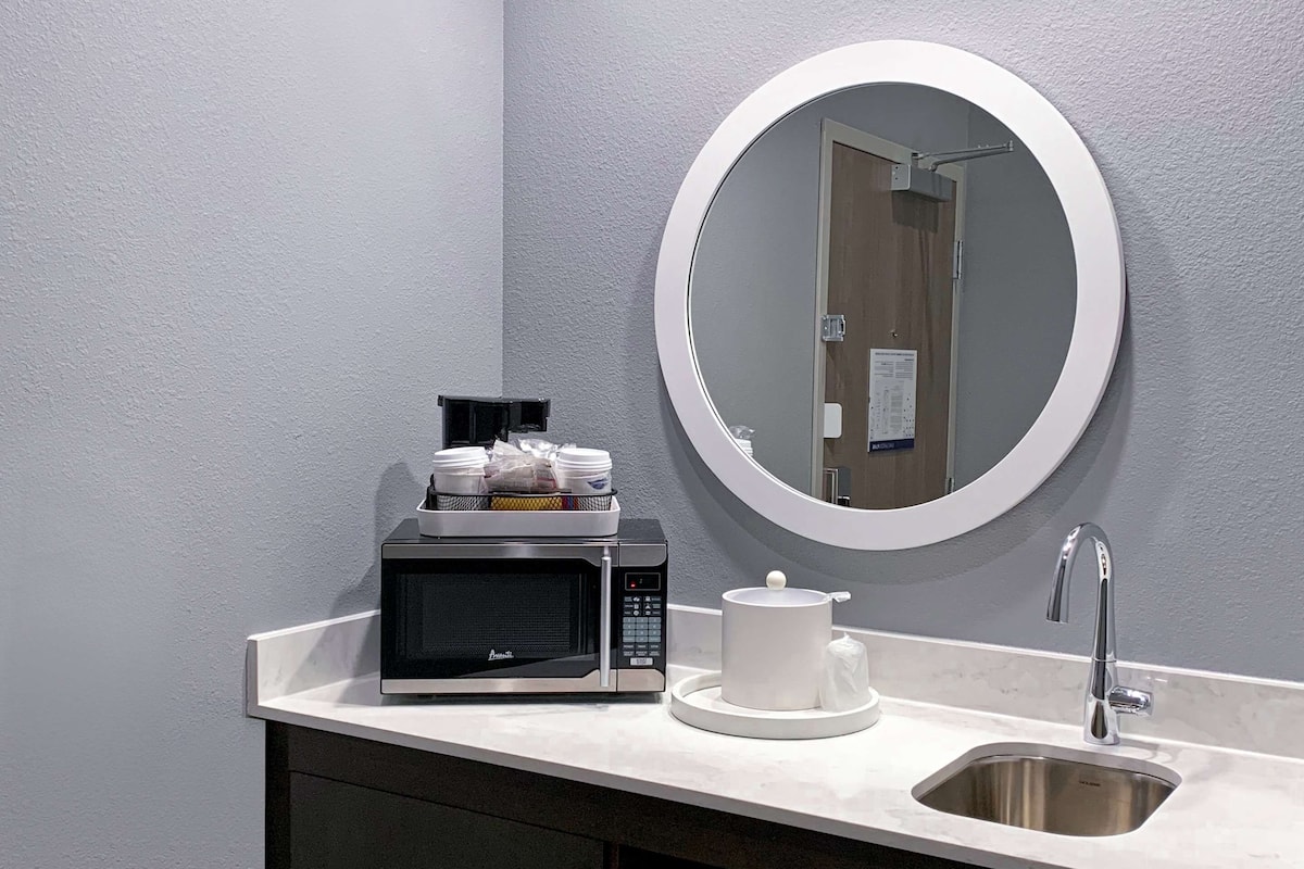 A modern bathroom with a white sink, a round mirror, a black microwave, and a small cabinet.