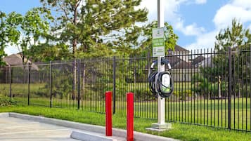 A parking area with electric vehicle charging stations, a fence, and greenery in the background.