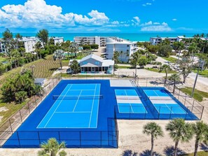 A tennis court complex with multiple courts, surrounded by buildings and palm trees, with a view of the ocean.