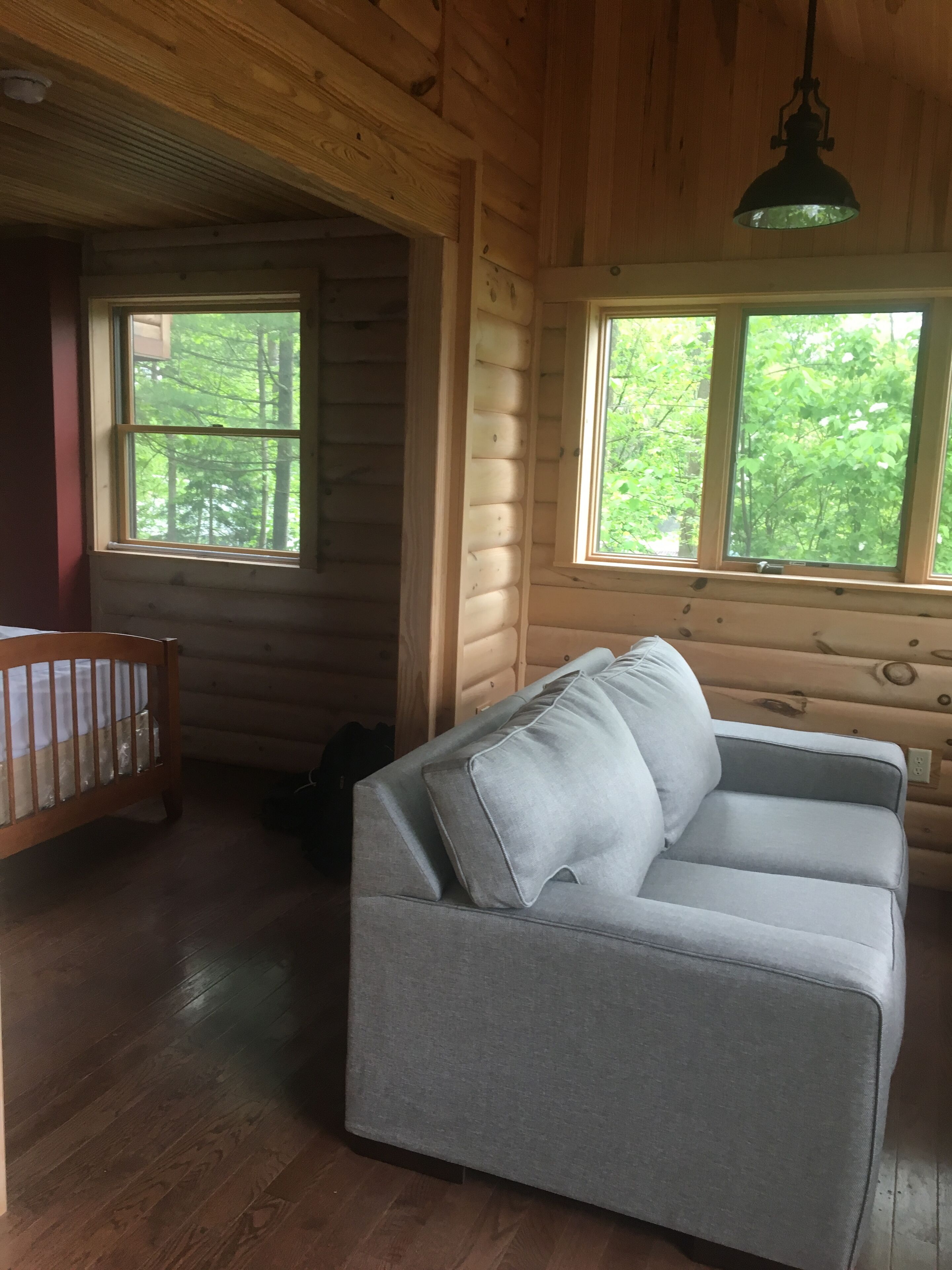 A log cabin interior with a grey sofa, wooden crib, and large windows offering a view of trees.