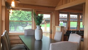 A wooden dining area with a table and chairs, a vase with plants, and large windows with a view of greenery. - Pointe of View on Otsego Lake. 4 Bedrooms, 4 Bathrooms, 6 miles from Cooperstown (Cooperstown)