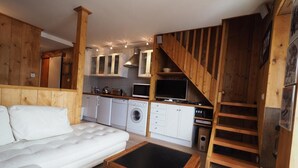 Un espacio habitable compacto con sofá, mesa de comedor, zona de cocina con electrodomésticos y una escalera de madera.