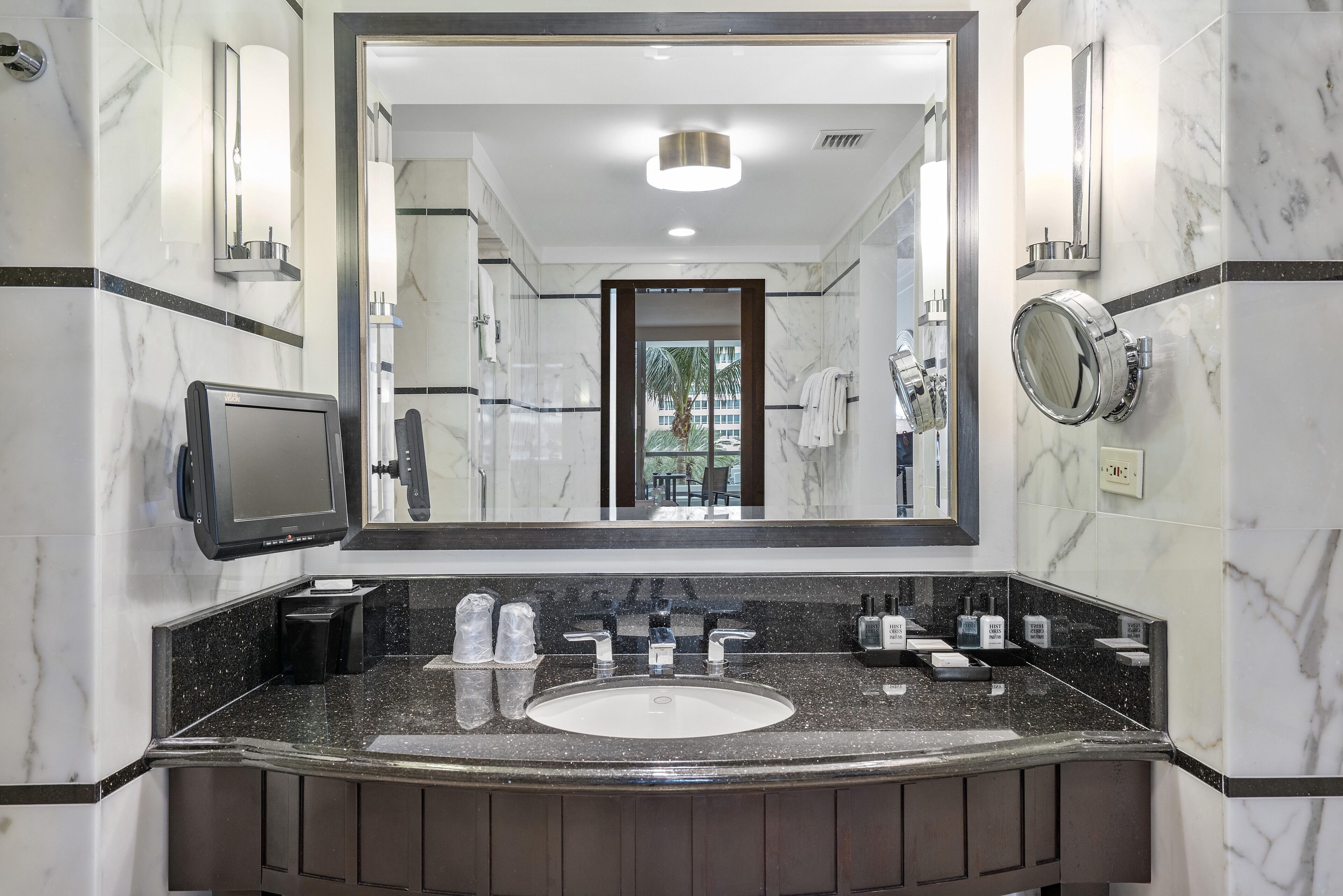 Un baño moderno con un espejo grande, paredes de mármol, un lavamanos con encimera negra y un inodoro con espejo redondo.