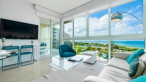 Ein modernes Wohnzimmer mit großem Fernseher, einem Essbereich mit Stühlen, einer Couch mit Kissen und Blick aufs Meer.