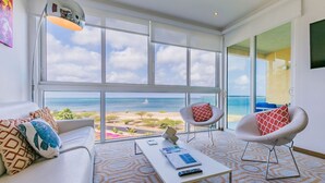 Un salon moderne doté d’une grande fenêtre donnant sur une plage, d’un canapé blanc, de deux fauteuils et d’une table basse avec des télécommandes.