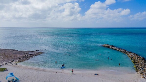 A beach with clear turquoise water, a rocky breakwater, and a sandy shore with a few people and a blue canopy.