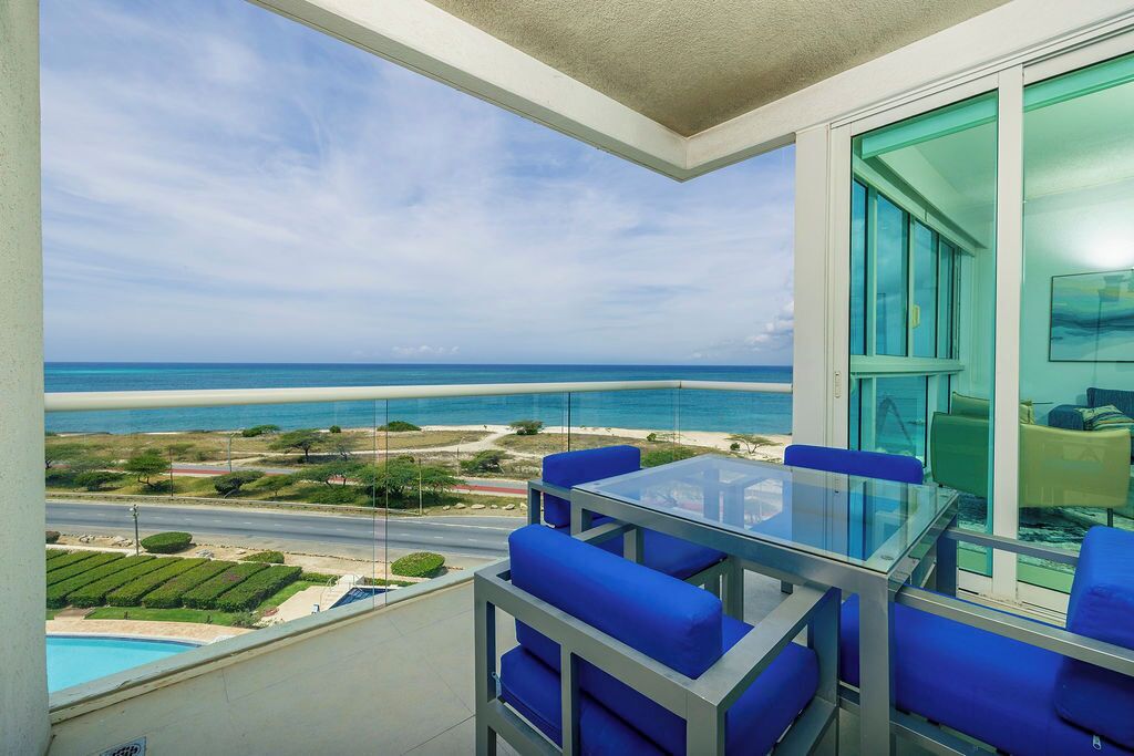 Un balcon avec une table en verre et des chaises bleues, donnant sur une plage et l’océan.