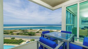 Un balcon avec une table en verre et des chaises bleues, donnant sur une plage et l’océan.