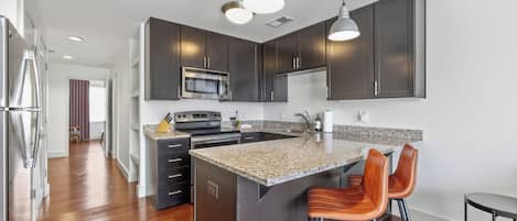 A modern kitchen with dark cabinetry, a central island with bar stools, stainless steel appliances, and pendant lighting.
