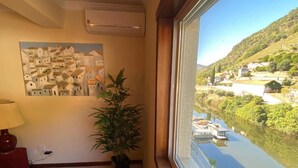 A room with a large window overlooking a river and a hillside village. There is a painting on the wall and a potted plant near the window. - Apt. in Pinhão, heart of Alto Douro wine region, vineyards and Douro River view (Pinhão)