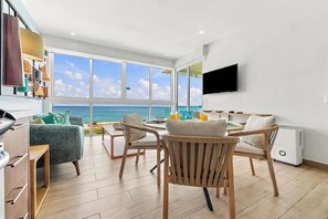 A modern living room with a large window overlooking the sea, a flat-screen TV mounted on the wall, and a dining area with wooden furniture.