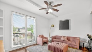 Una sala moderna con sofá, reposapiés y sillón, ventilador de techo, lámpara de pie y un amplio ventanal con persianas.