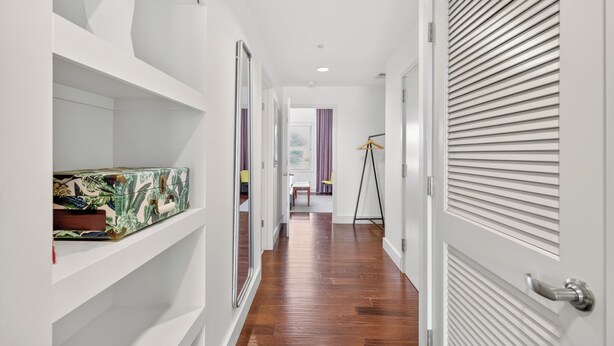 Un pasillo con piso de madera, paredes blancas y una puerta cerrada con persianas horizontales.