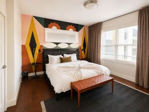 A hotel room with a bed, a bench, a wall-mounted TV, a window with curtains, and a patterned rug.