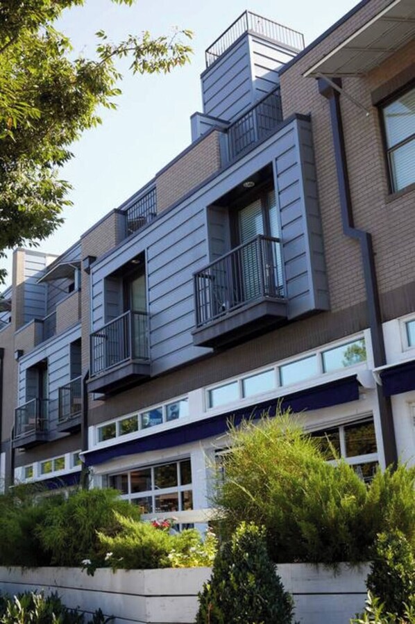 A modern multi-story residential building with balconies, large windows, and a landscaped front garden.