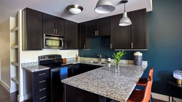 Une cuisine moderne avec des meubles de couleur sombre, des plans de travail en granit, un îlot central doté de tabourets, un four à micro-ondes, une cuisinière et des luminaires suspendus.