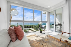 Enjoy a panoramic ocean and greenery view from this spacious living room with a sofa, coffee table, and wicker chair.